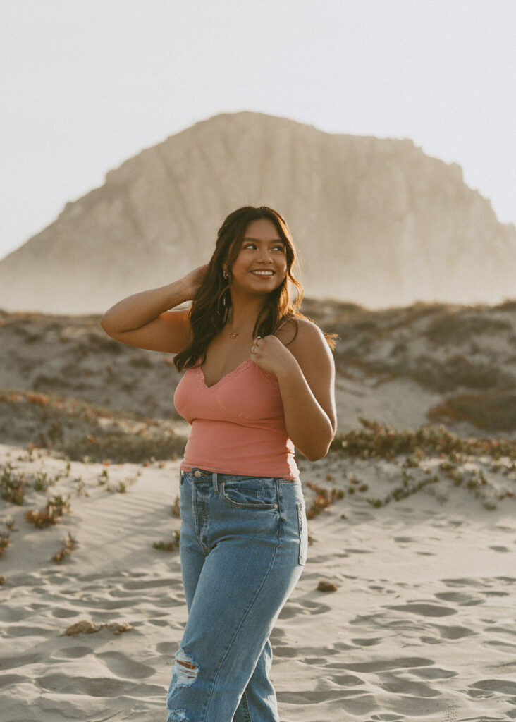 Woman wearing a pink tank top and blue jeans standing on a sandy beach, smiling and looking to the side