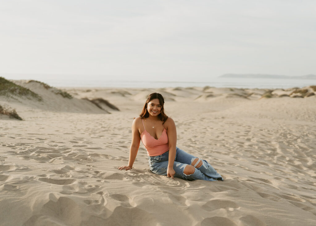 Woman sitting on sandy dunes smiling, wearing a pink tank top and ripped blue jeans