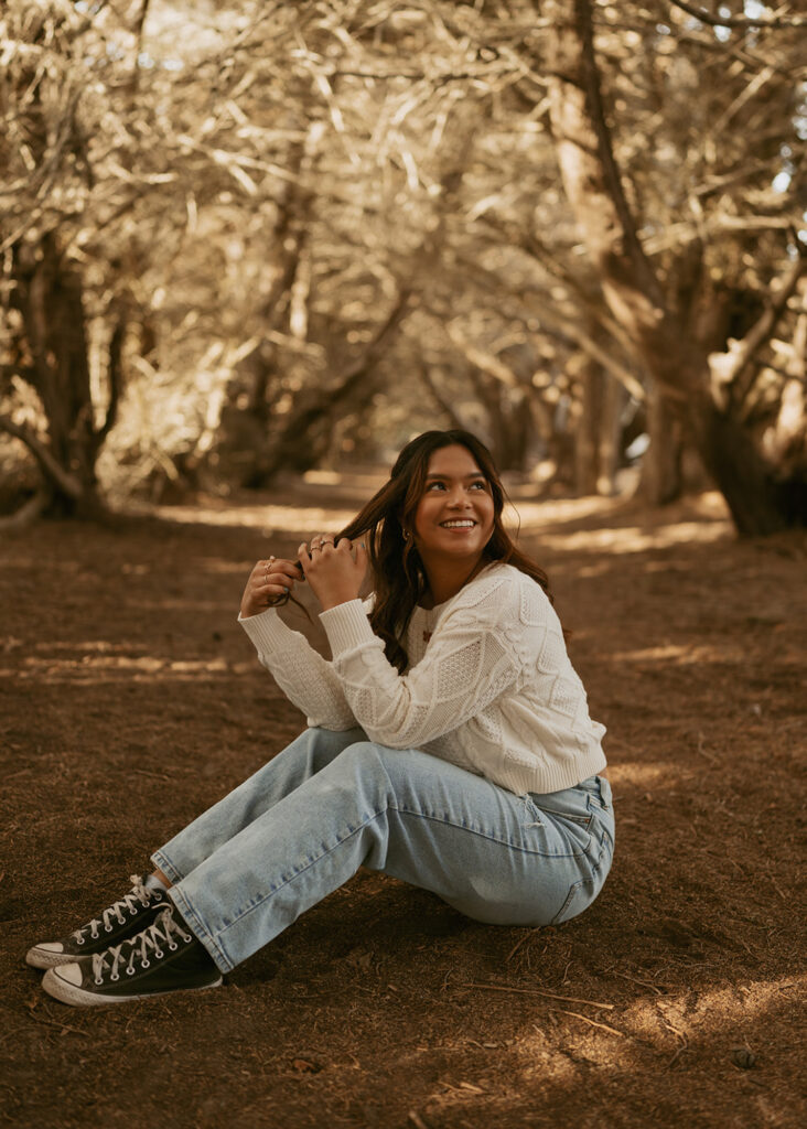 Woman in a white sweater and blue jeans sits on the ground in a forest, smiling and looking off to the side