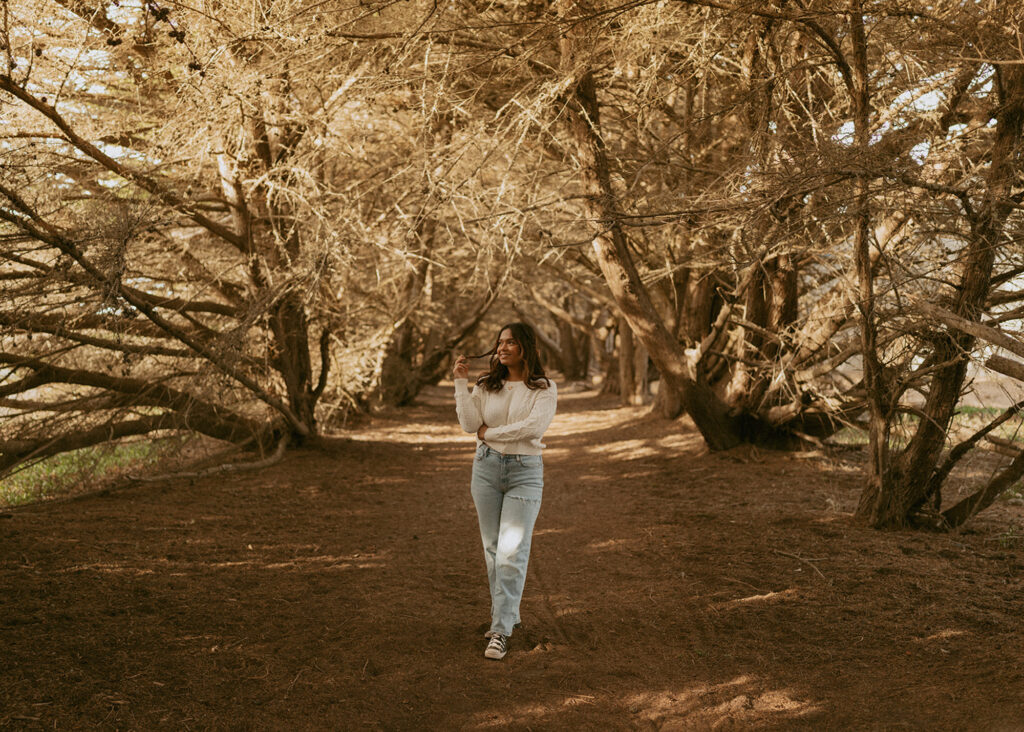 Woman walking along a sun-dappled dirt path surrounded by trees with intertwining branches