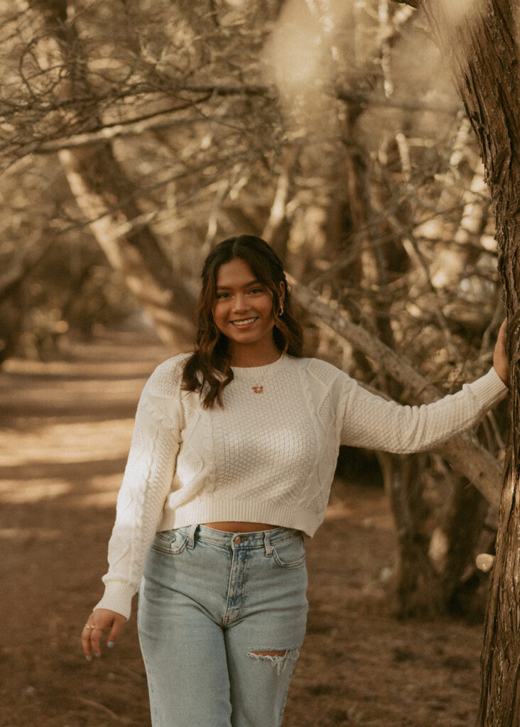 woman with long hair, wearing a white sweater and blue jeans leaning against a tree with one arm while looking at the camera and smiling