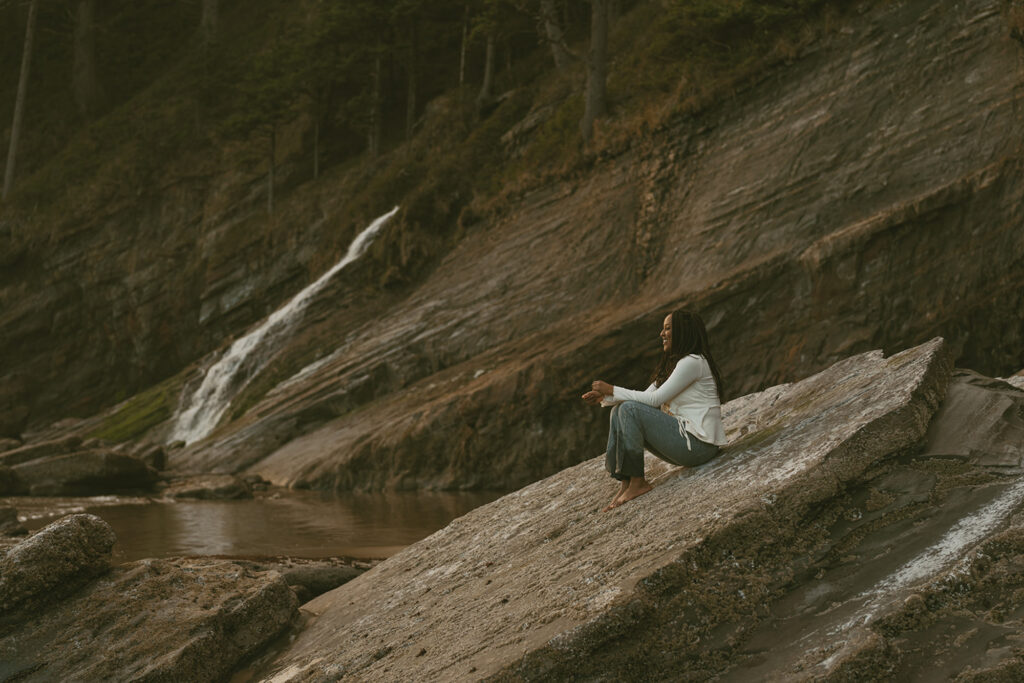 Woman with long hair sits barefoot on a large rock near a flowing waterfall 