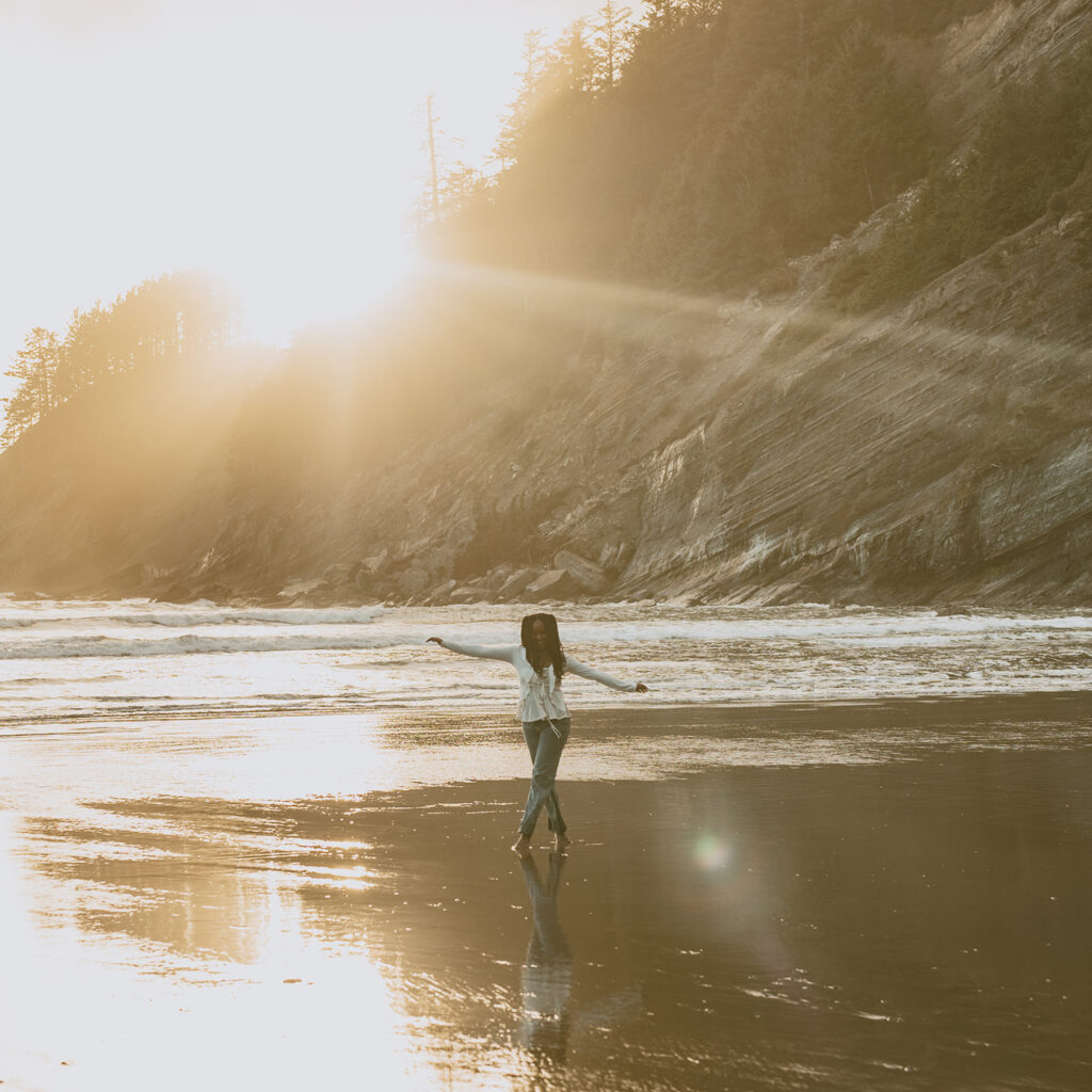Woman standing on a beach, facing the ocean with arms outstretched, as the sun sets behind a cliff covered in trees