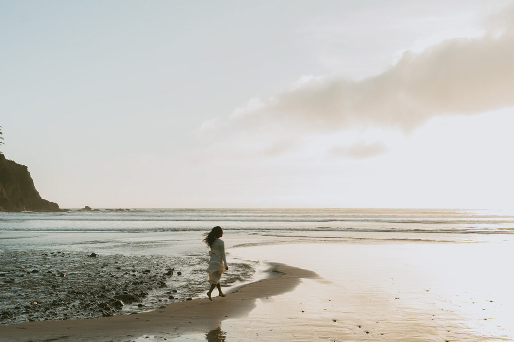 Woman wearing a white dress running along a sandy beach towards the ocean as the sun sets