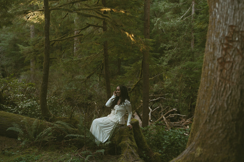 Woman in a long white dress sitting on a large fallen tree trunk amidst a dense, sunlit forest