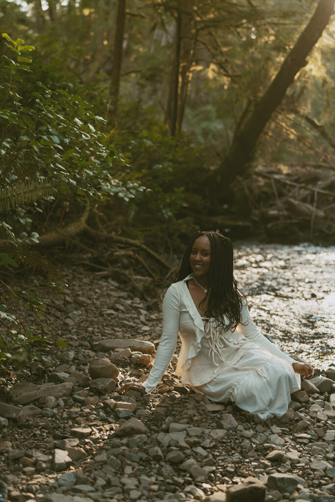 Woman with long braided hair sitting on a rocky riverbank, wearing a white dress surrounded by trees and greenery, with sunlight filtering through the leaves