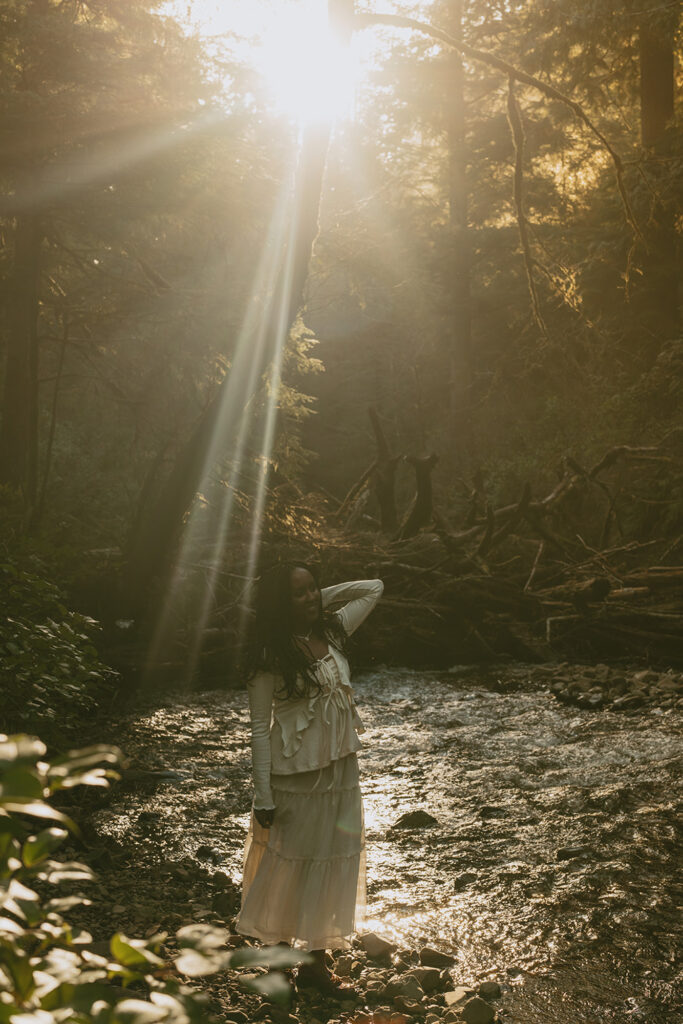 Woman standing in a shallow stream within a forest, with sunlight streaming through the trees