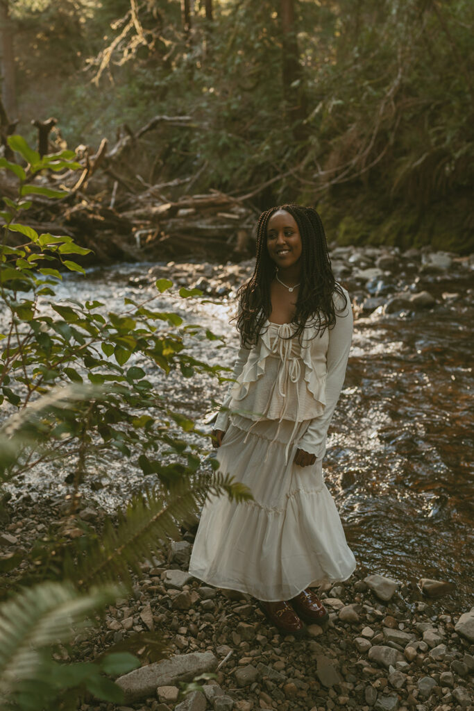 Woman with long braided hair, wearing a flowing white dress, standing on a rocky riverbank