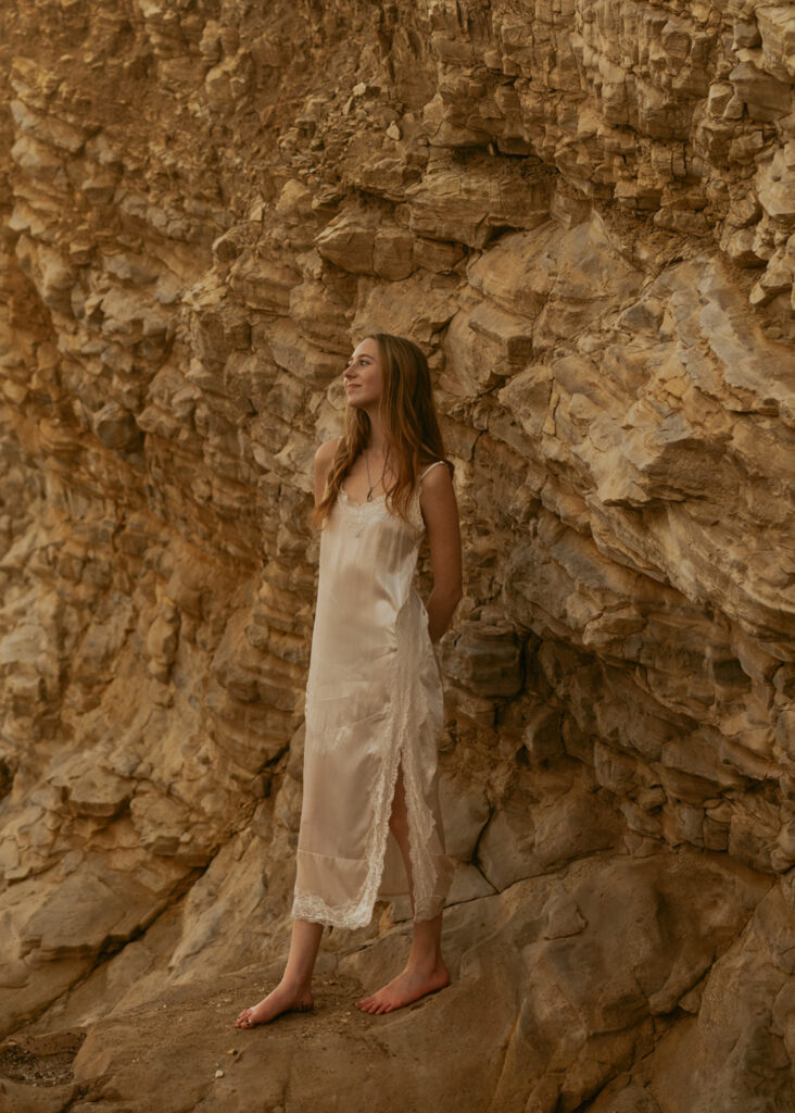 Woman in a white dress stands barefoot against a rugged rock formation