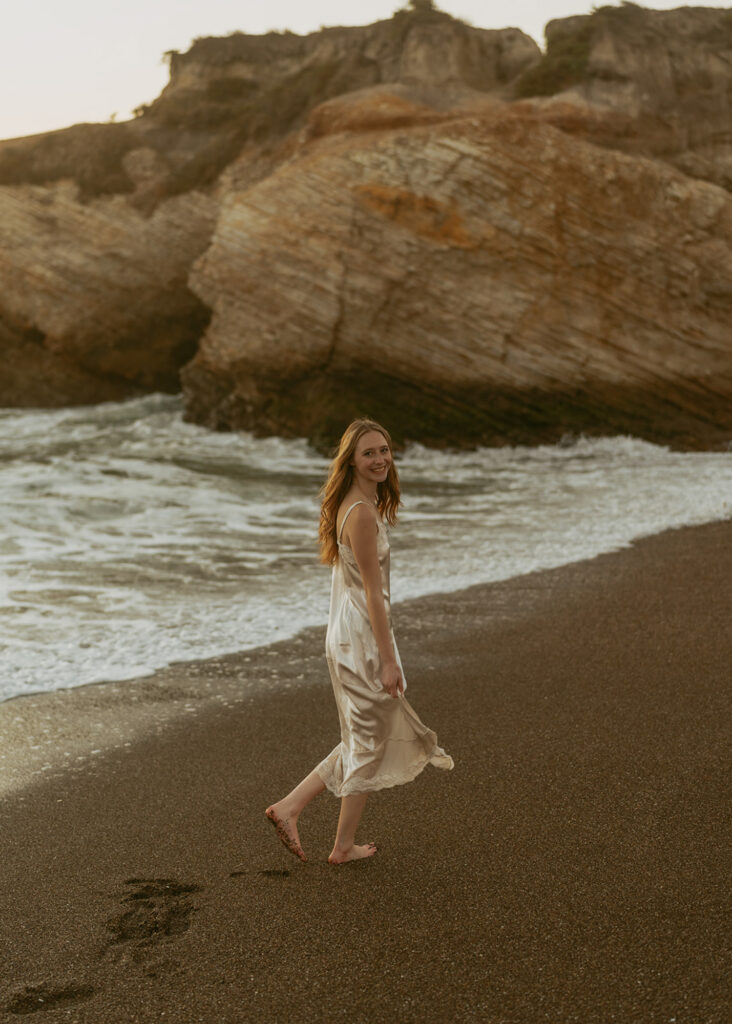 Woman in a flowing white dress walks barefoot along the beach, smiling and looking back towards the camera for a beautiful senior beach photo
