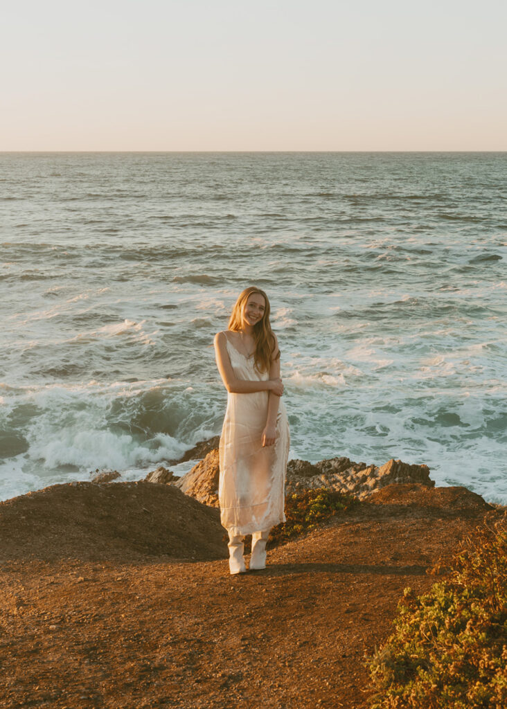 Woman in a light dress stands on a rocky cliff overlooking the ocean