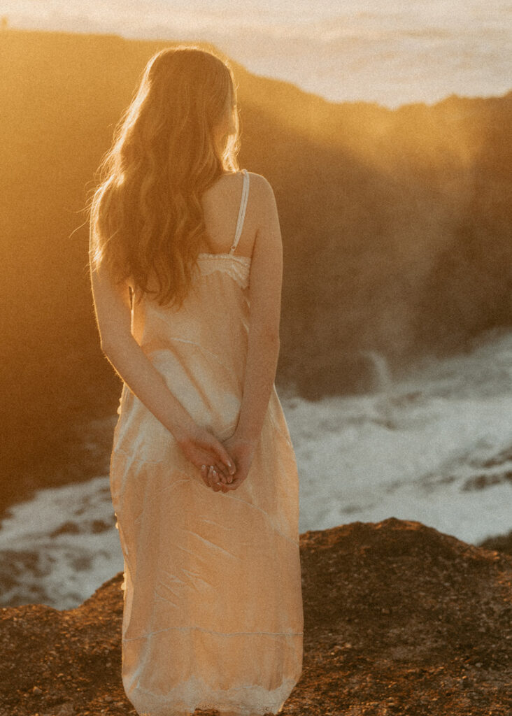Woman wearing a light, flowing dress, hands clasped behind her back, with long, wavy hair stands on a rocky shore, facing the ocean as the sun sets