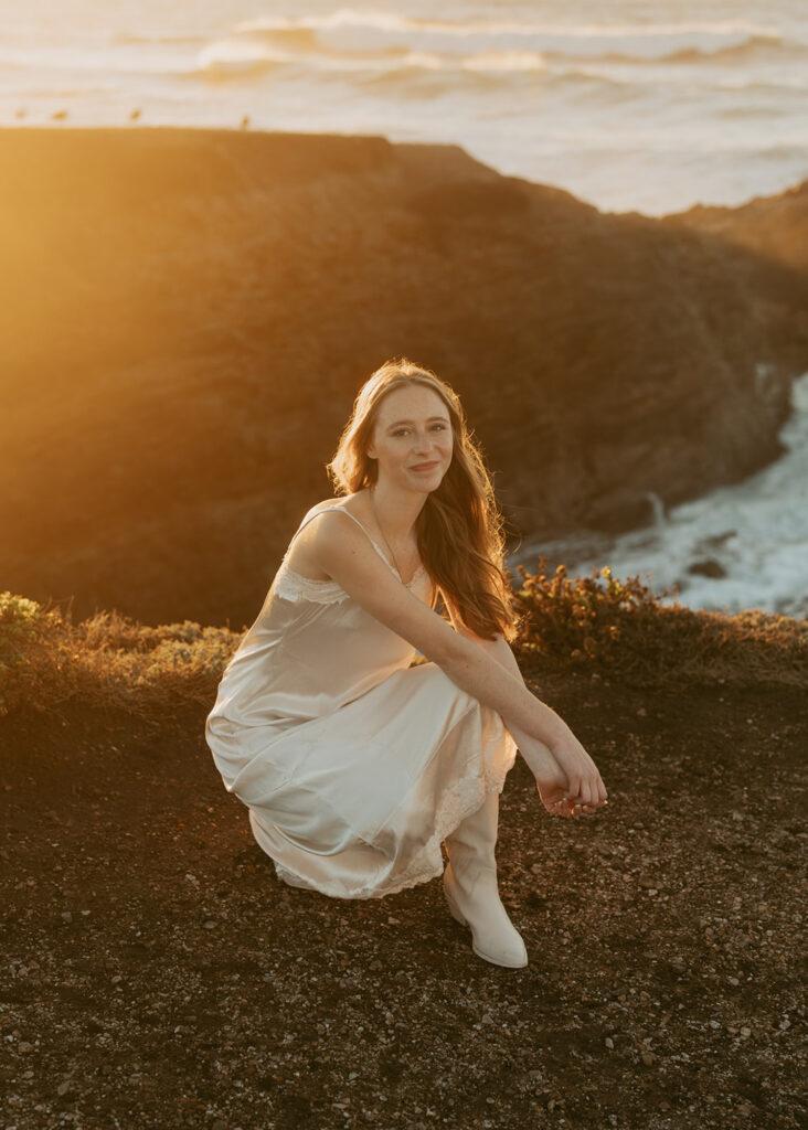 Woman in a flowing white dress crouches on a rocky cliff by the ocean, bathed in warm, golden light during sunset