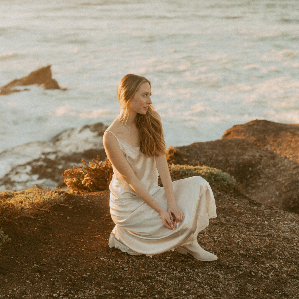 Woman in a white dress sits on a rocky ledge by the ocean at sunset for her Senior beach photo
