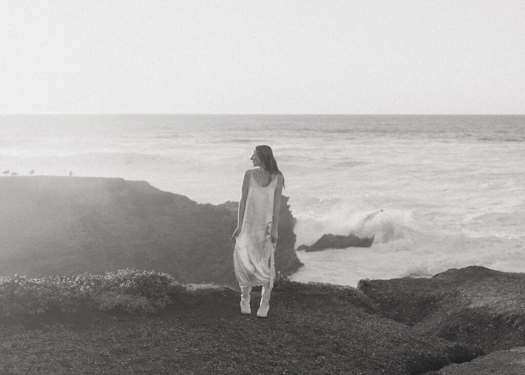 Woman stands on a rocky cliff overlooking the ocean for her senior beach photo