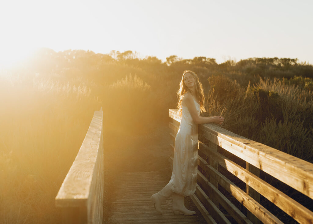 Lady wearing a white dress leans against a wooden railing on a sunlit boardwalk surrounded by tall grass and foliage