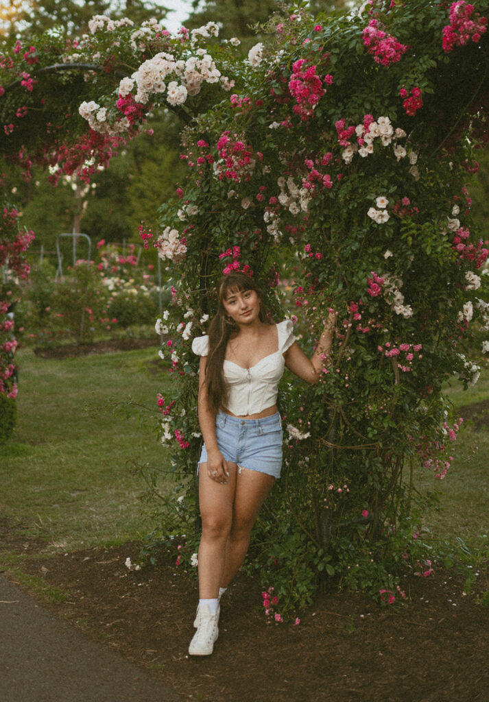 Woman standing in a Rose Garden in Portland wearing a white crop top and denim short for her Senior Pictures
