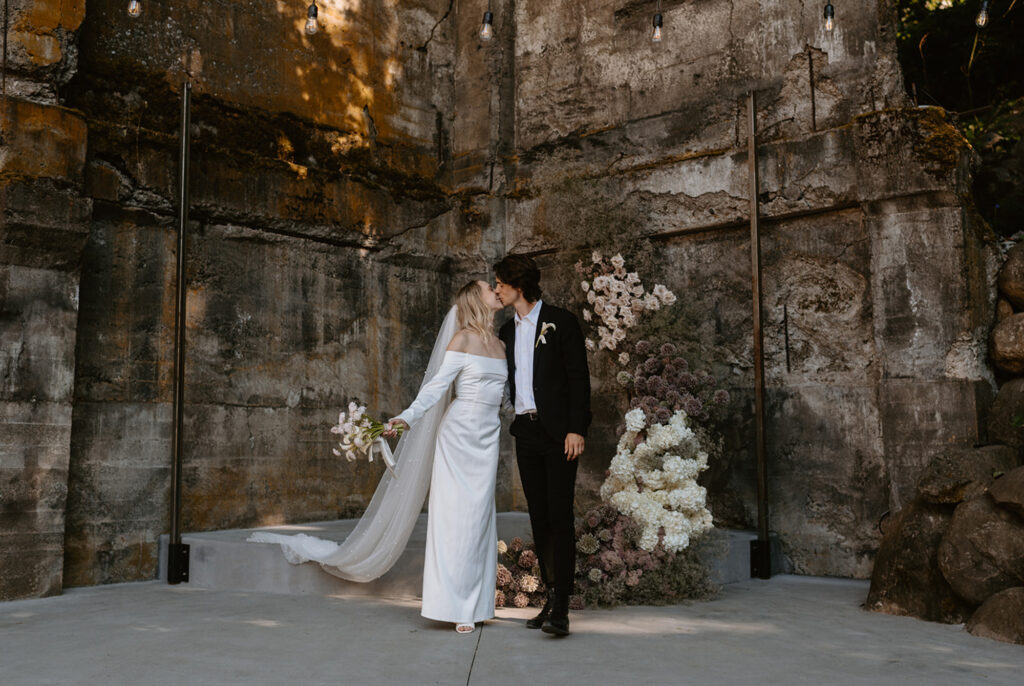 Couple kisses in front of a minimalistic floral backdrop, highlighting the beauty of an intimate elopement ceremony.