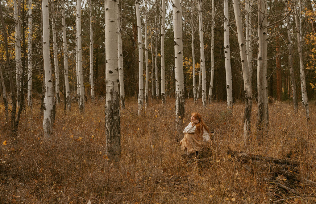 Senior girl with long red hair sitting on a fallen log in a golden autumn aspen grove in Bend, Oregon