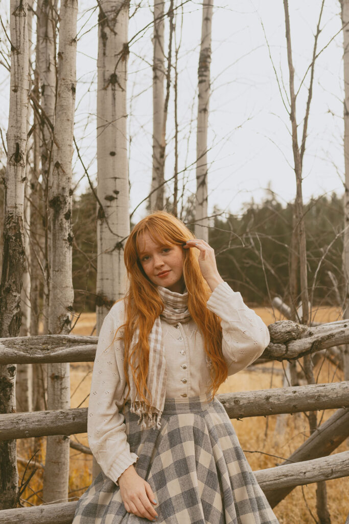 Senior girl with long red hair resting her hand on a rustic wooden fence, surrounded by aspens