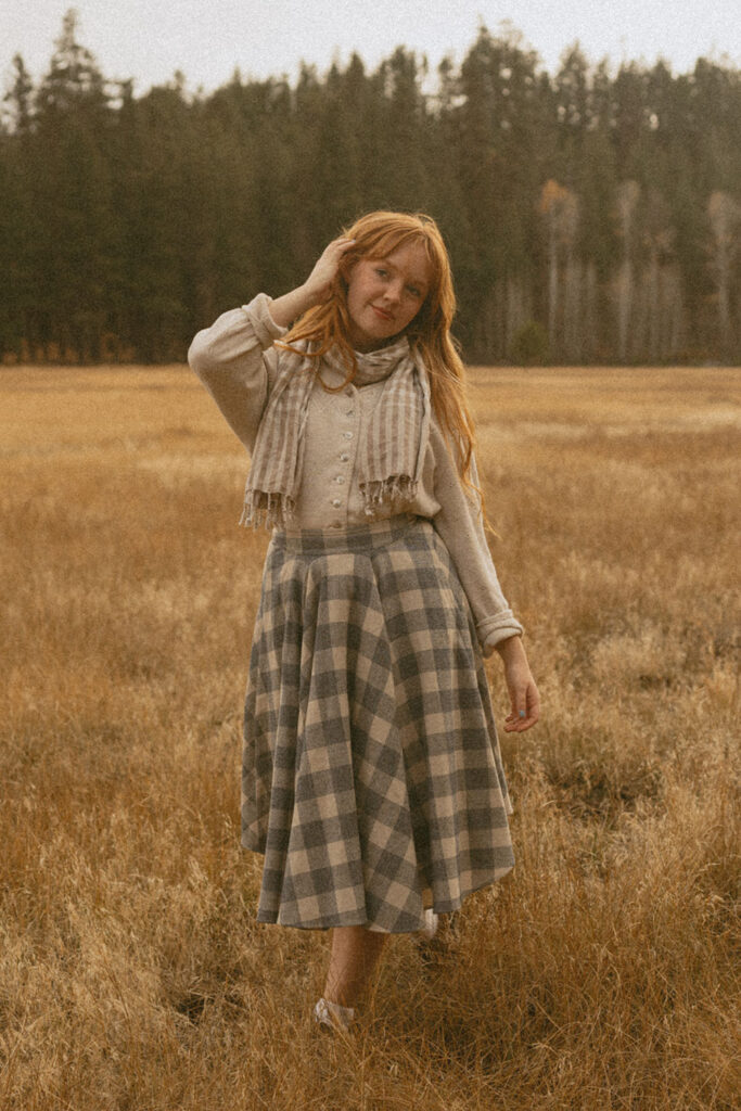 Senior girl smiling in an open field, adjusting her hair, wearing a vintage-inspired outfit