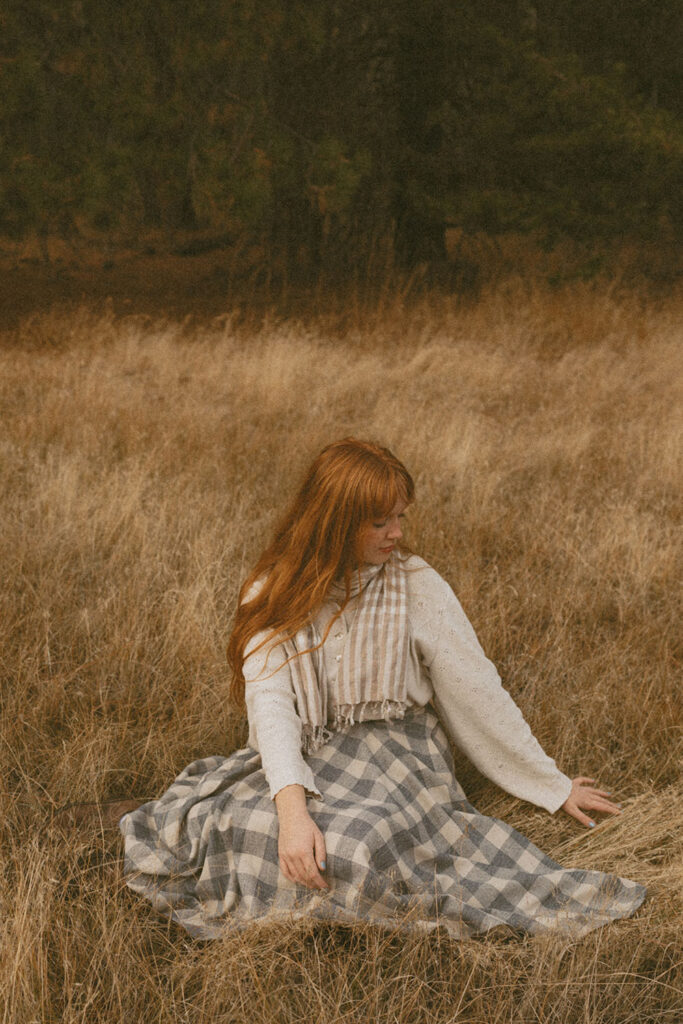 Senior girl sitting in a field, gently touching the grass for a photo session by a senior photographer in Bend