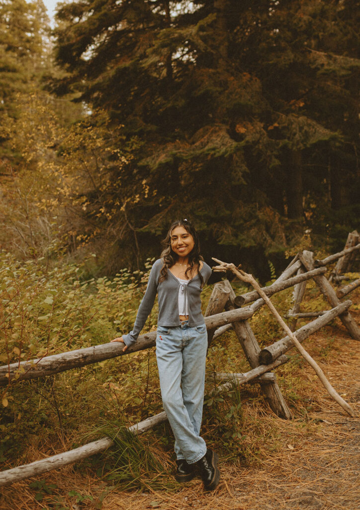 Senior girl in jeans and a cardigan, casually leaning on a wooden fence in the woods