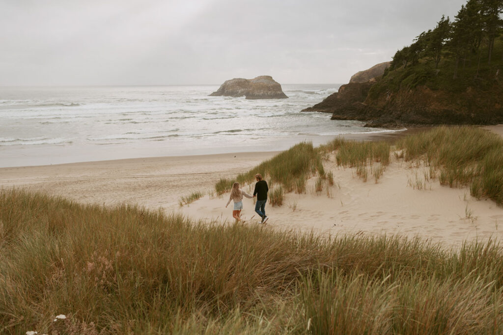 Callie and Nolan walking hand in hand with the view of Haystack rock behind them