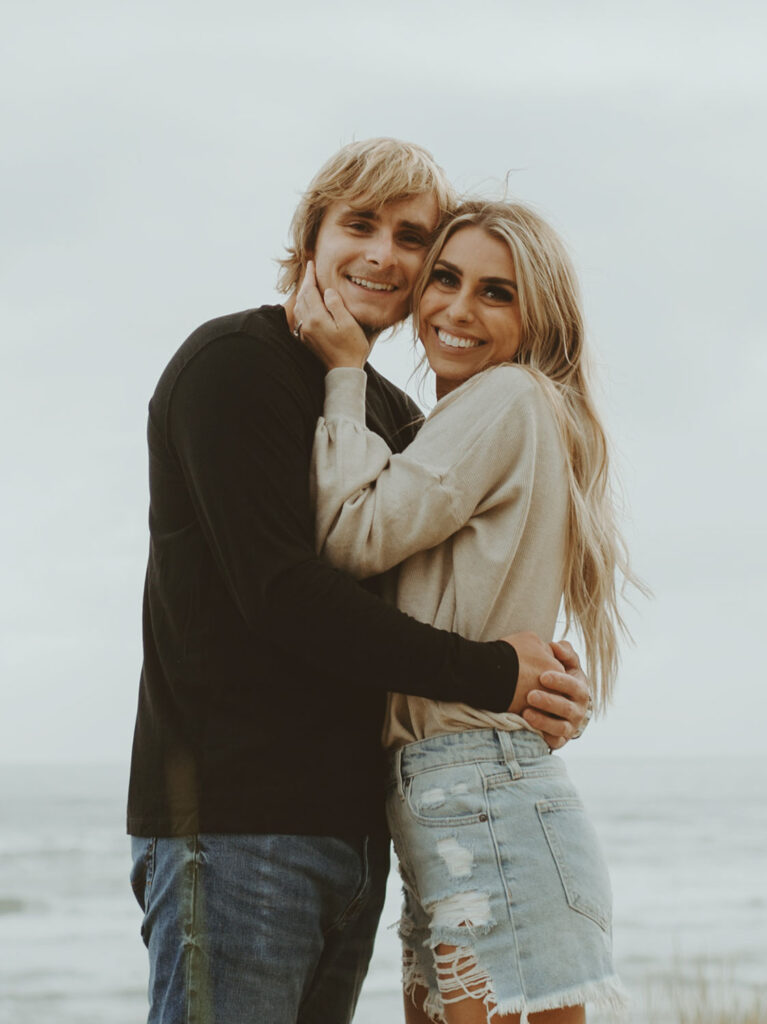 Callie and Nolan stands closely at the beach, smiling and embracing