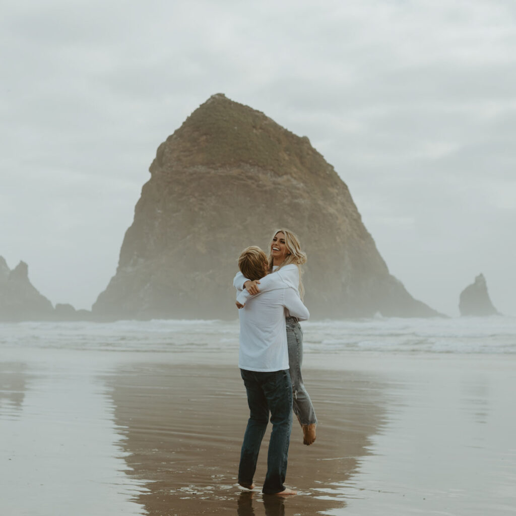 Nolan lifts Callie off the ground as she laughs, the ocean and a towering sea stack in the background