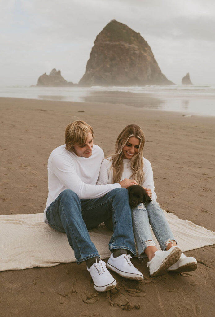 Callie and Nolan sit on the beach with their black puppy, gently holding and looking at it with love