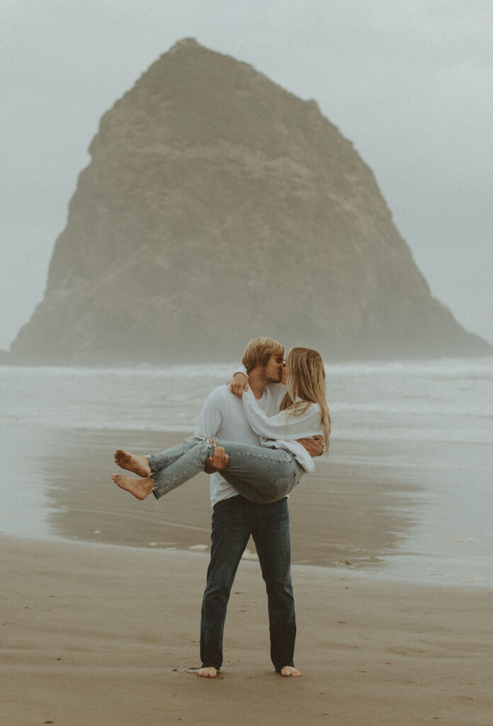 Nolan holds Callie in his arms, her legs playfully kicked up as they share a romantic kiss on the misty beach