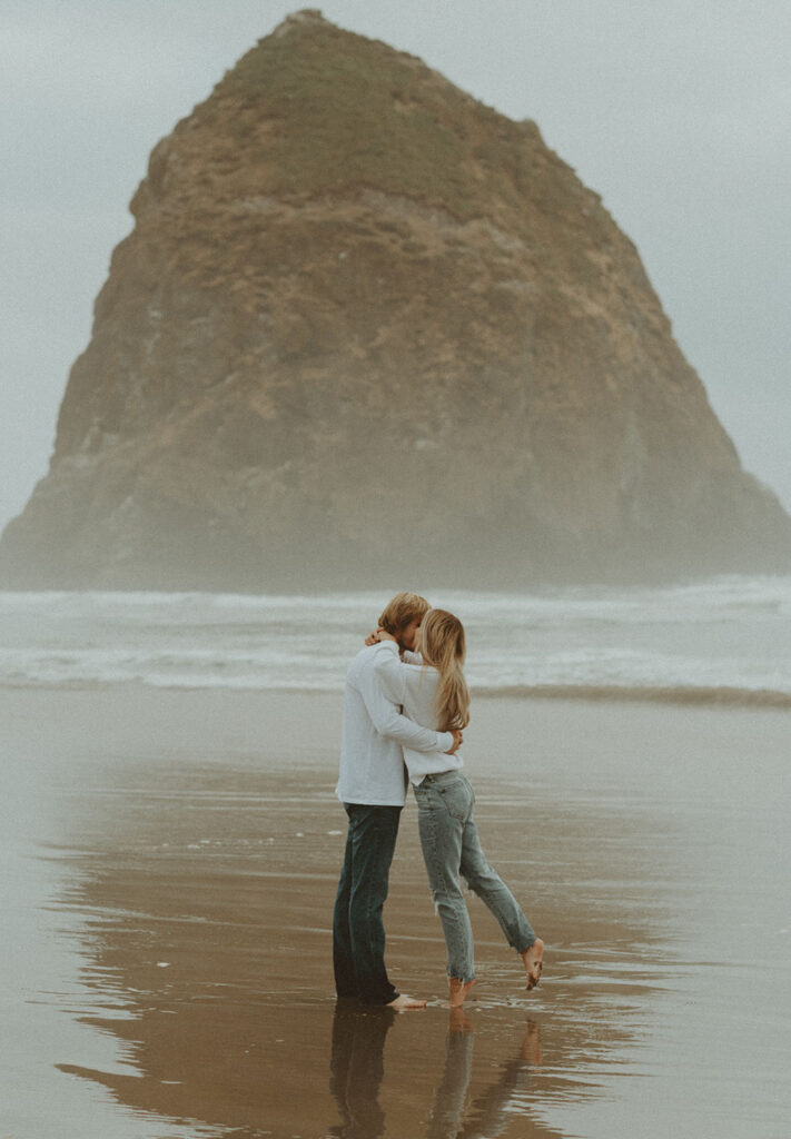 Callie and Nolan embrace on the wet sand, sharing a kiss as their feet sink slightly into the water