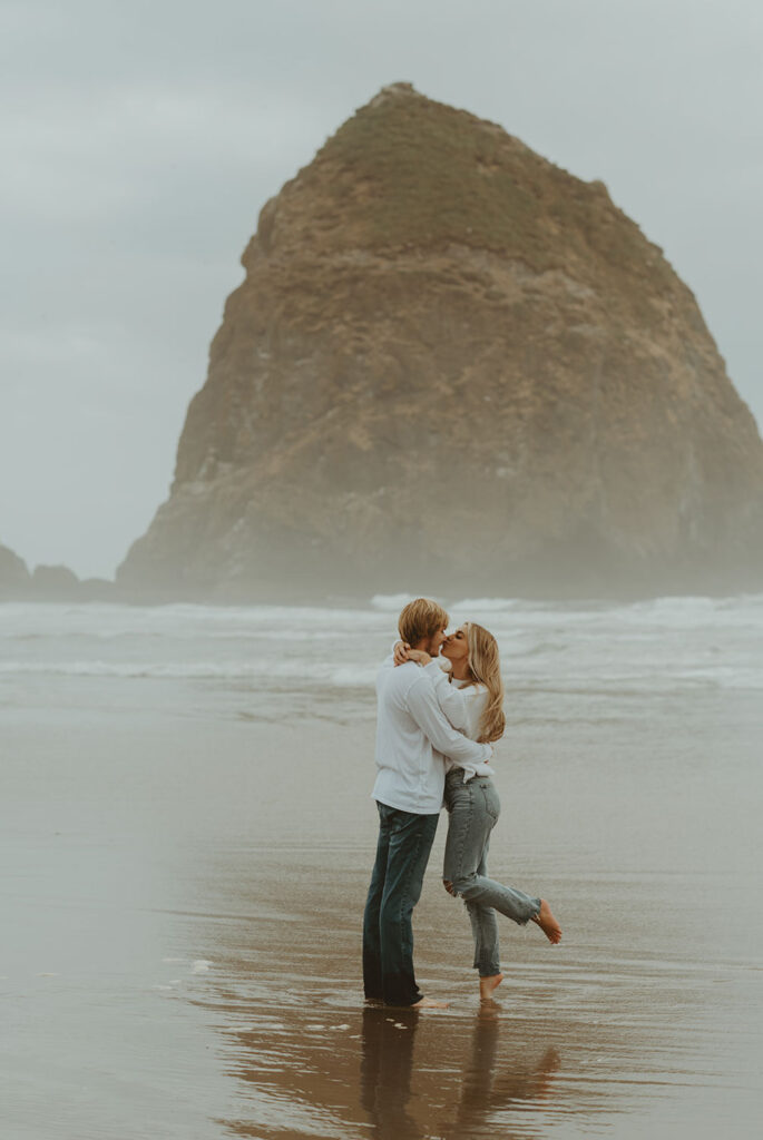 Callie and Nolan stand barefoot in the shallow water, holding each other close and kissing in front of the sea stack