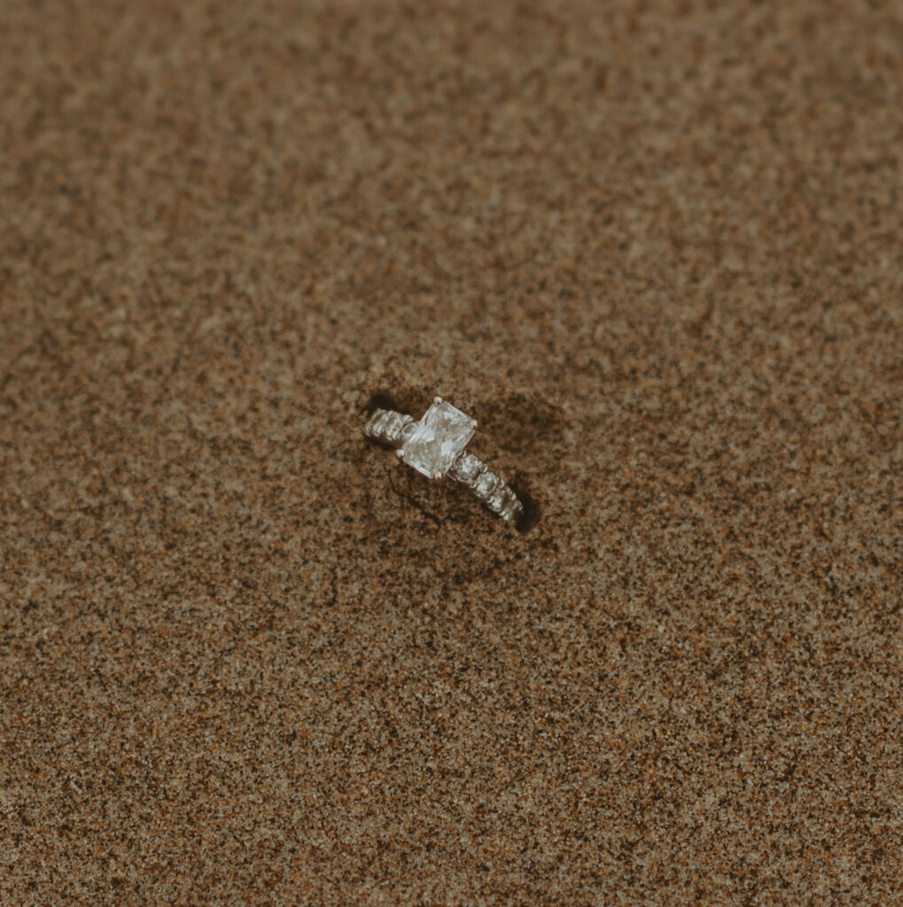 A radiant engagement ring sits in the sand, sparkling against the earthy tones