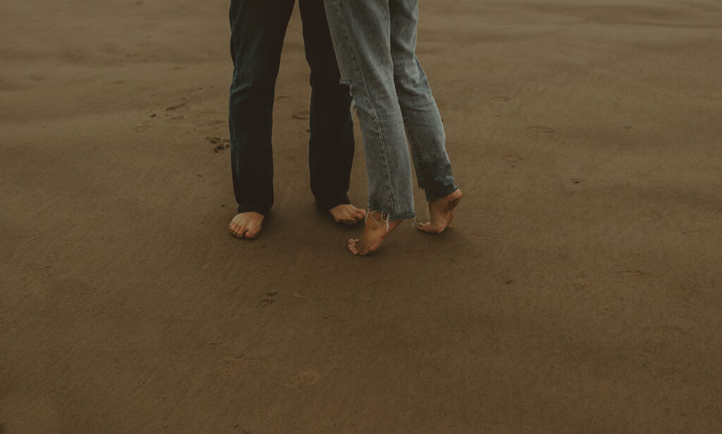 Callie and Nolan stand barefoot on the wet sand, feet close together