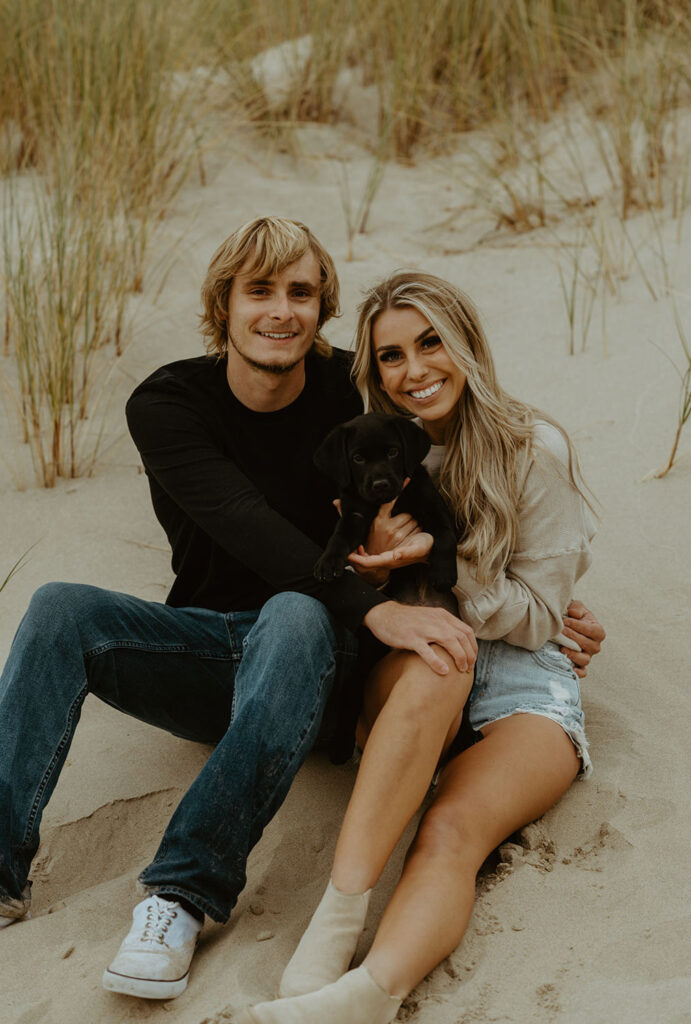 Callie and Nolan sit together on the sand, holding their black puppy and smiling at the camera