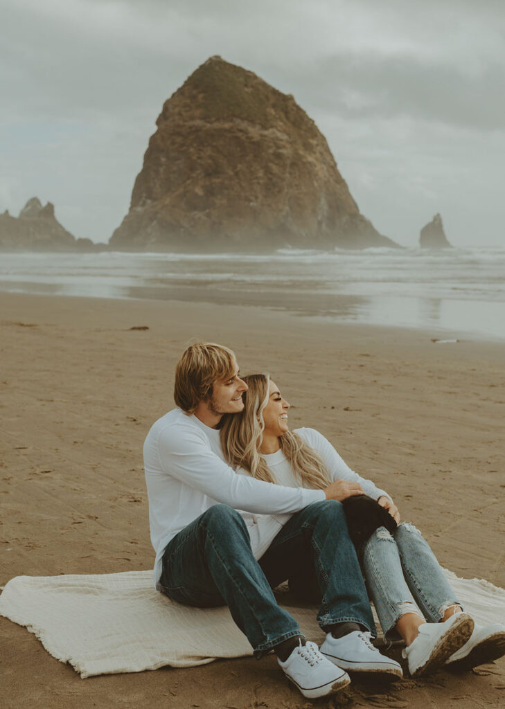 Callie and Nolan sits on a blanket on a sandy beach, embracing and smiling at the ocean