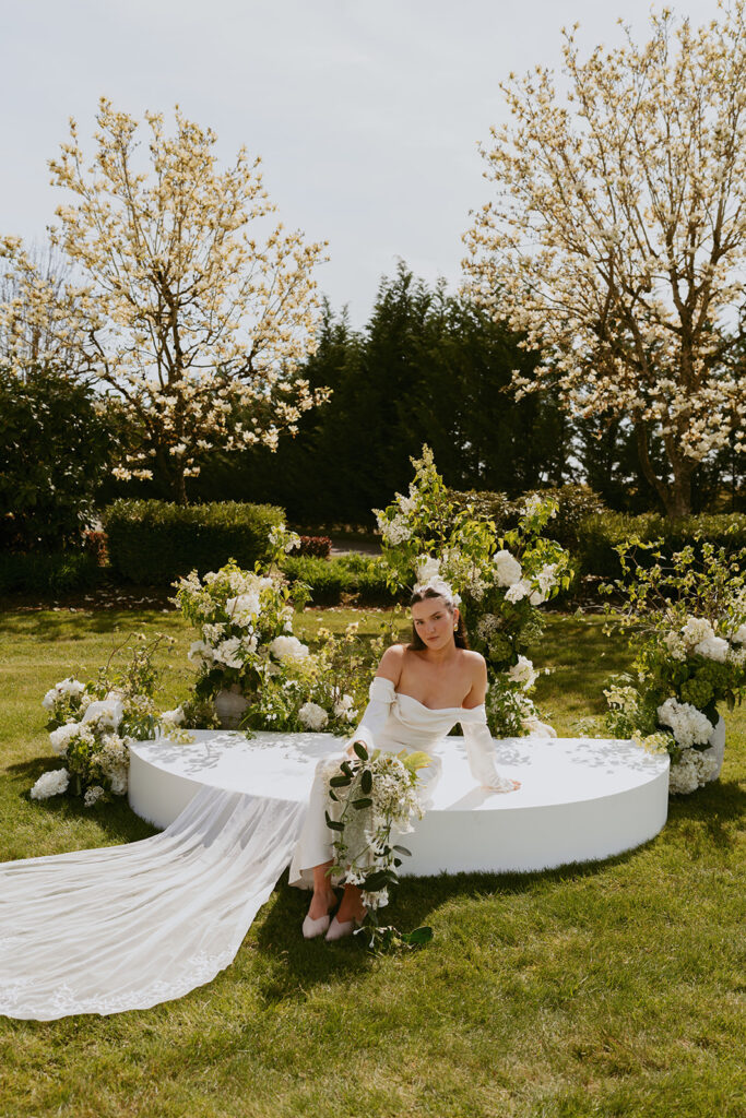 A bride in a white dress is sitting on a white platform in the grass.