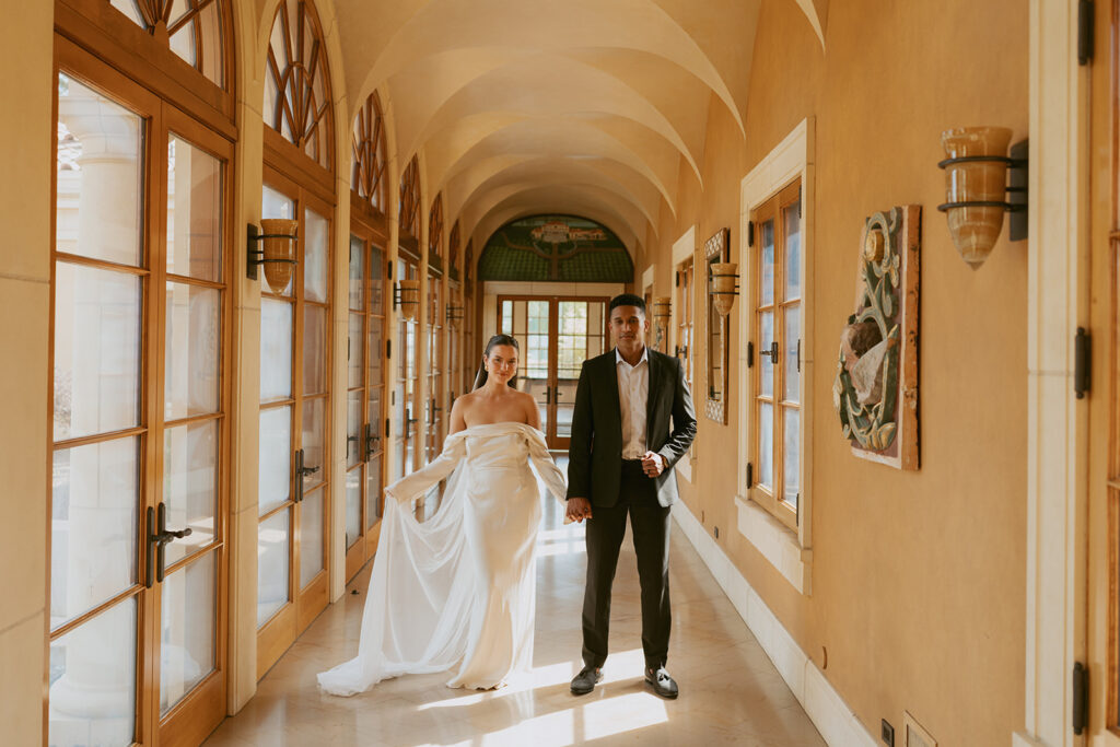 A bride and groom are walking down a hallway holding hands.