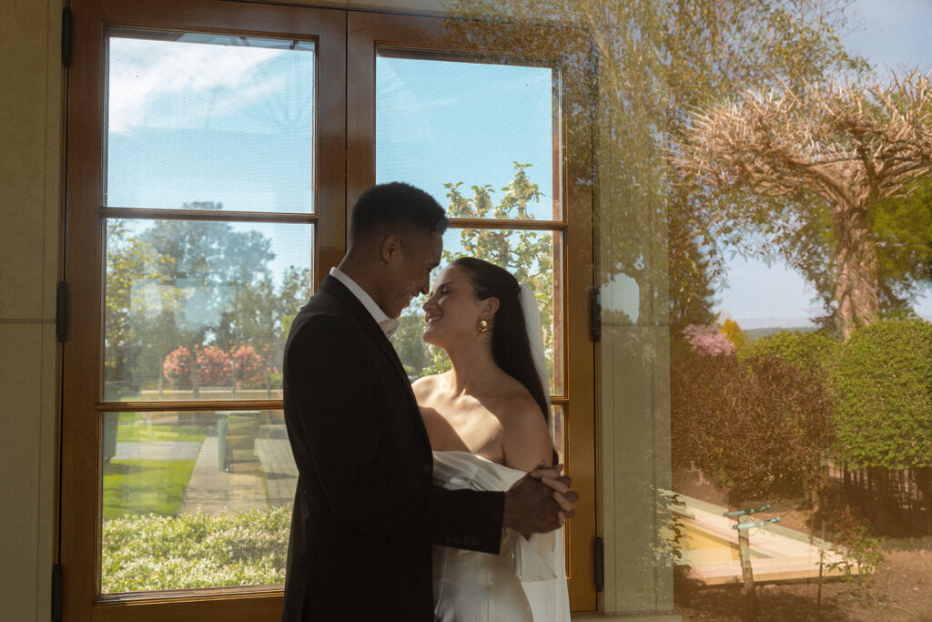 A bride and groom are dancing in front of a window.