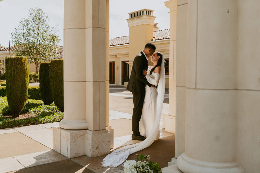 A bride and groom are kissing under a pillar in front of a building.