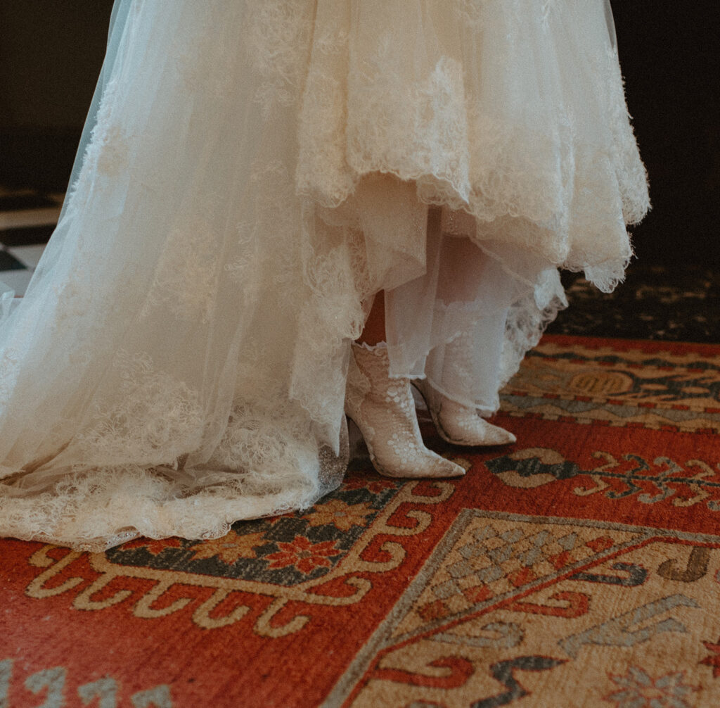 A bride in her wedding dress and cowboy boots is standing on a carpet