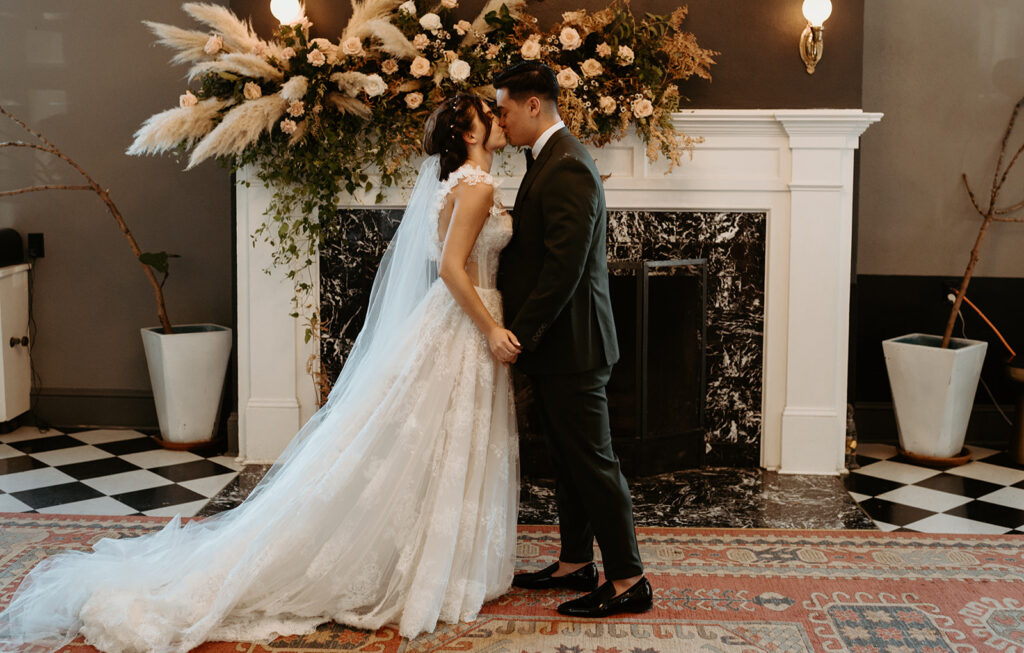 A bride and groom are kissing in front of a fireplace.