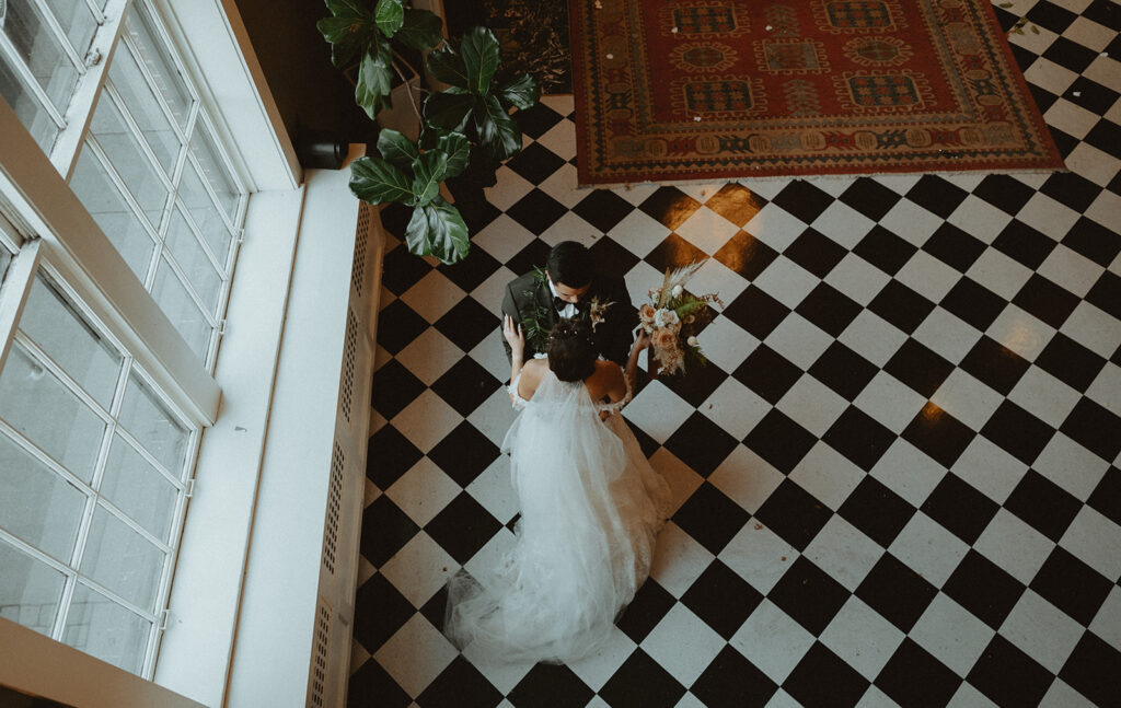 Bride and groom standing on a checkered floor next to a window.