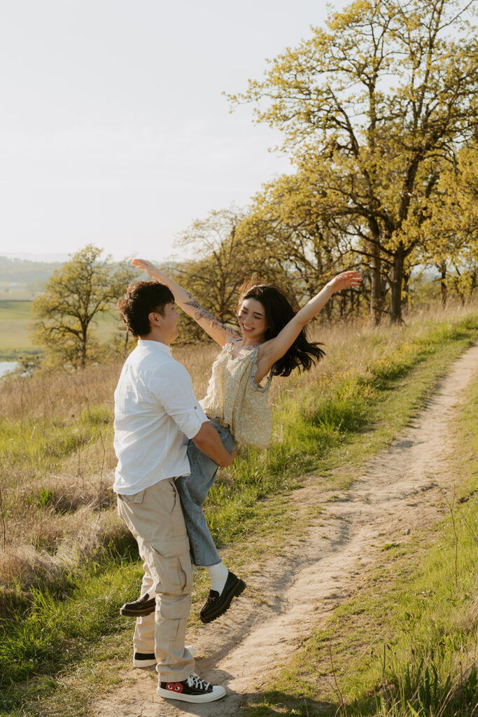 Man lifting his fiancée as she throws her arms up joyfully on a nature trail