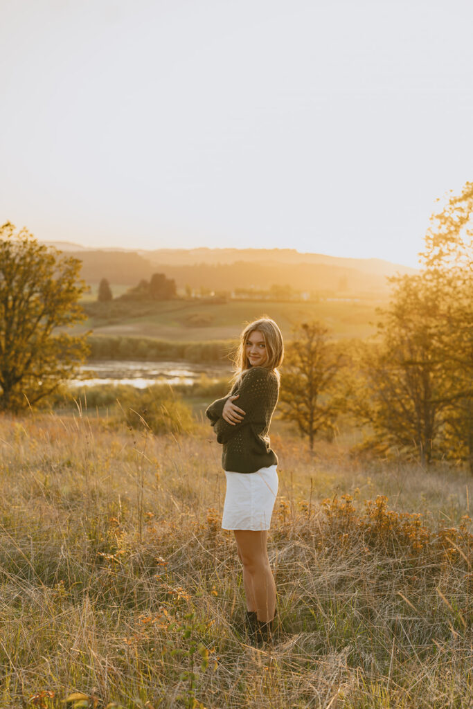 Young woman standing in a field at sunset, arms crossed and smiling