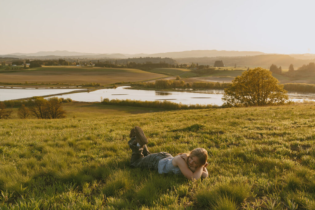Wide view of a woman laying on a hilltop field with a lake and sunset behind