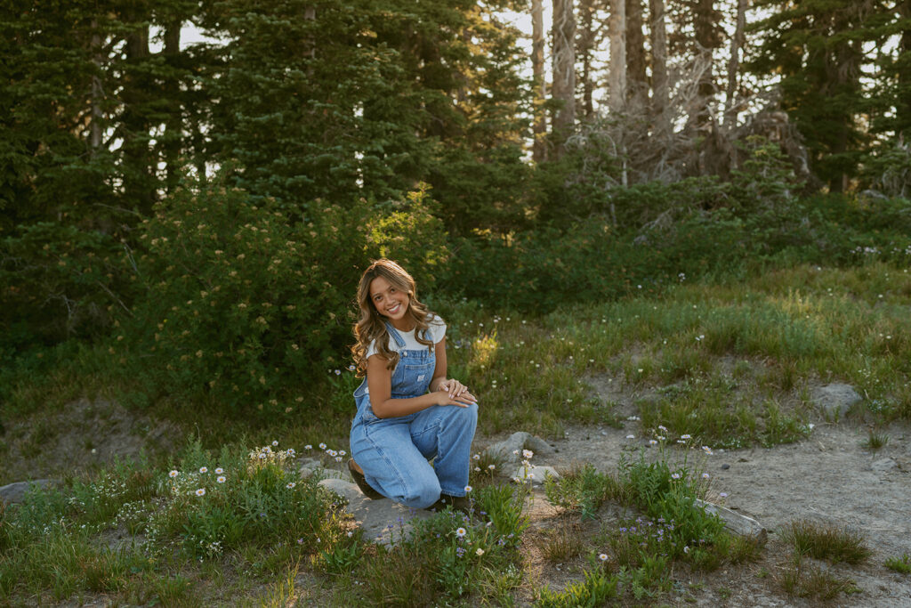 A woman in overalls is kneeling down in a field in the woods.