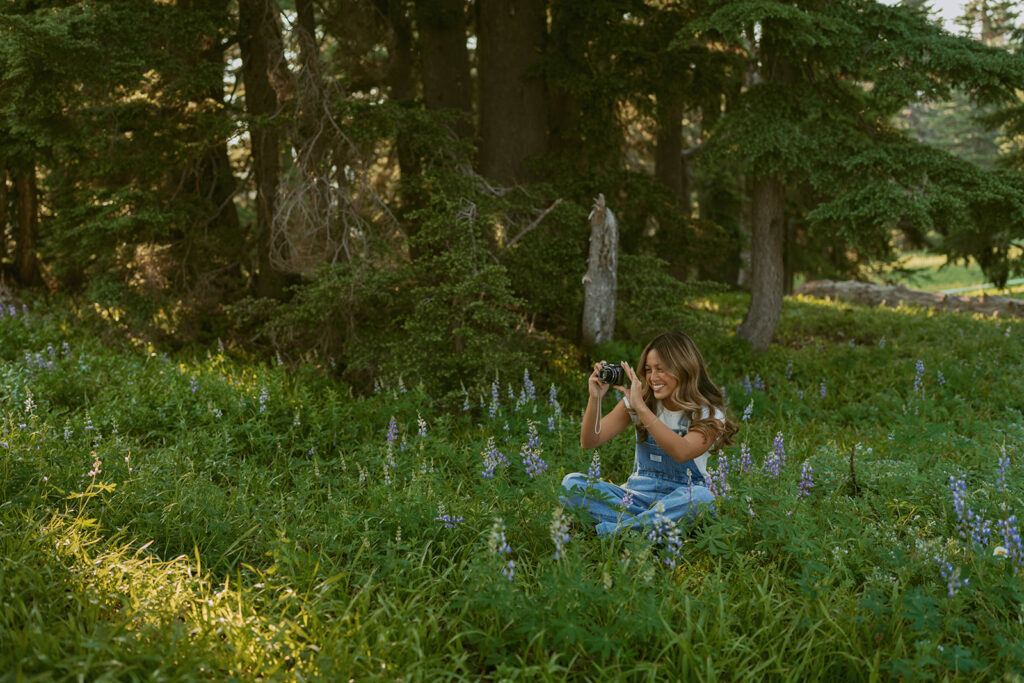 A woman is sitting in a field of flowers taking a picture with a camera.